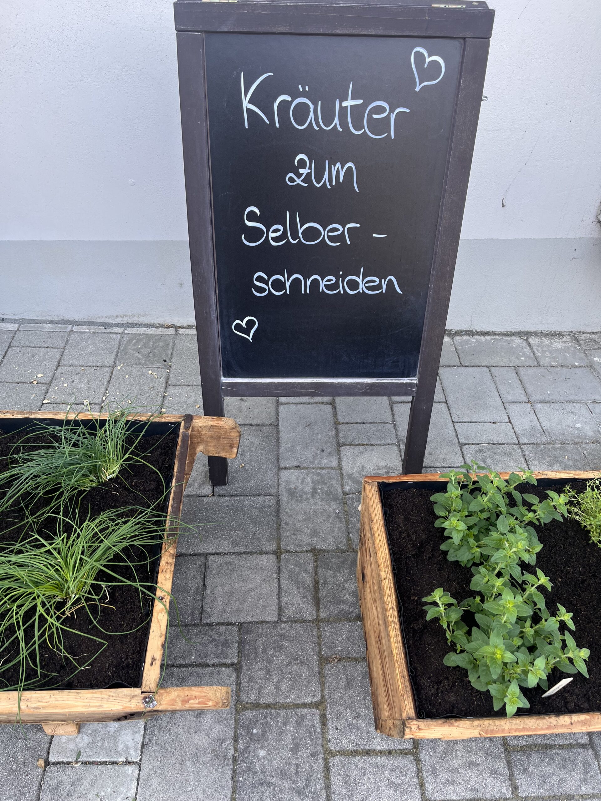 In dem Bild sieht man ein Schild, auf dem in Kreide steht: "Kräuter zum Selberschneiden" mit zwei Herzen. Im Vordergrund sieht man den Anfang zweier Holztröge mit Kräutern.